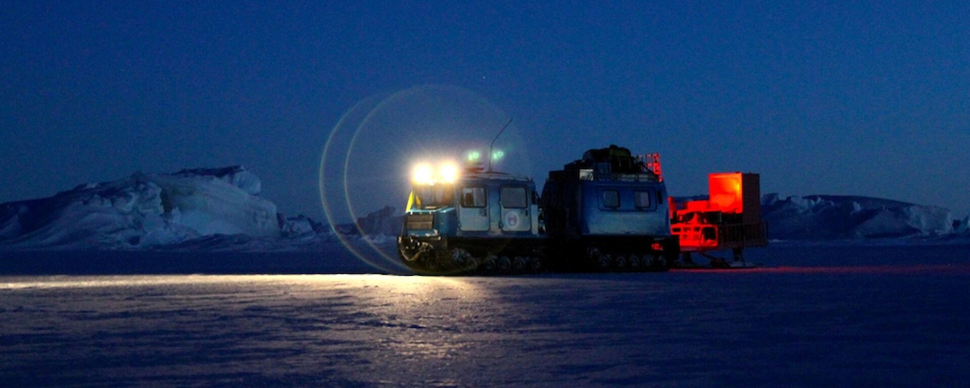 The blue Hägglunds vehicle towing a sled with it headlights on in the early hours of the morning