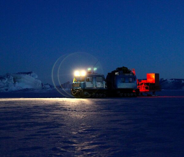 The blue Hägglunds vehicle towing a sled with it headlights on in the early hours of the morning