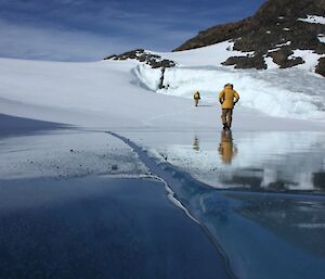 Two expeditioners walking on a snow drift that ends on a frozen fresh water lake