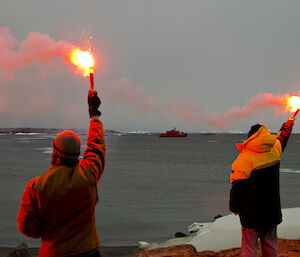 2 expeditioners waving goodbye with lit flares facing the ship as it departs