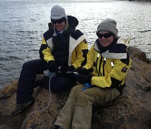 Two expeditioners sitting on a boulder alongside a lake sampling