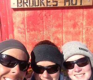 head and shoulder photo of 3 females with the hut in the background