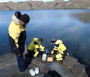 2 expeditioners sampling water at waters edge, 1 expeditioner looking on
