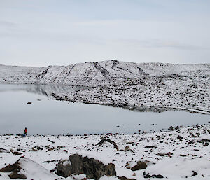 expeditioner standing at the end of a lake, plenty of snow cover around the hills