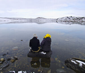 two expeditioners sitting on a rock on the end of a lake