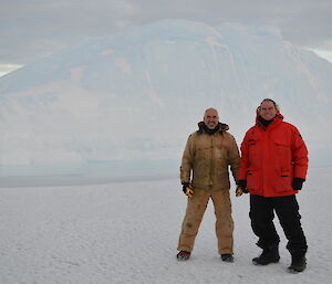 Two expeditioners standing on the sea ice with a large iceberg in the background