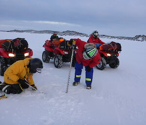 One expeditioner holds a drill whilst another drops a tape measure into hole