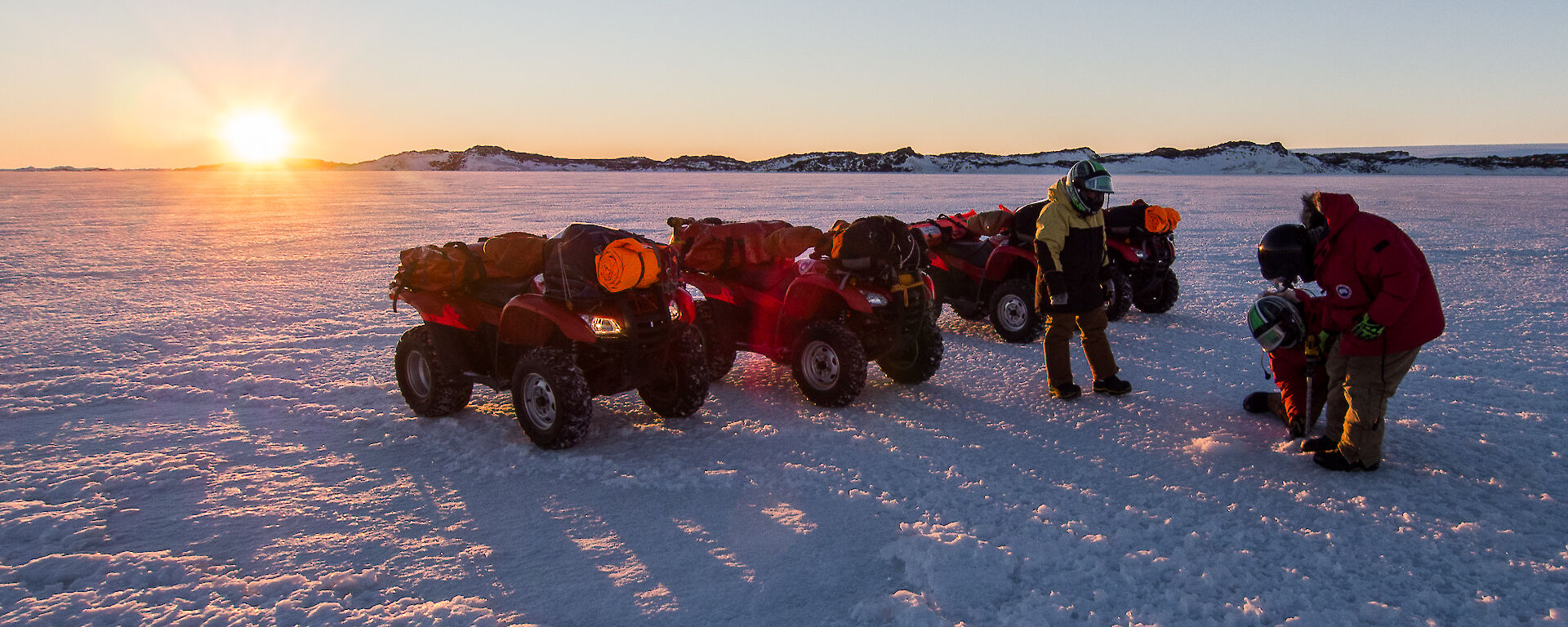Three people standing next to quads drilling sea ice