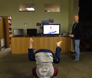 Expeditioner preparing to stand on his head in front of TV playing yoga class