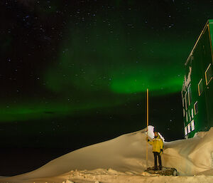 Expeditioner stands alongside the green main building with a green aurora overhead