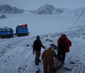 Three expeditioners looking at a small generator on the ice ground. Hagglund in the background.