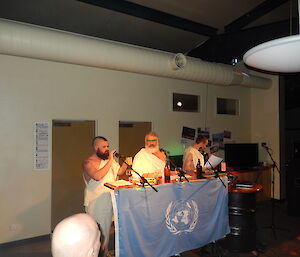 Three men in togas standing behind table with DJ equipment and microphones