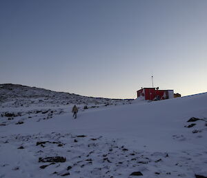 Expeditioner on snow walking towards a small red hut