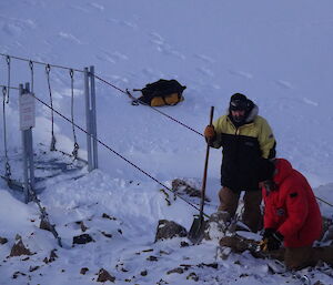 Two expeditioners with tools next to a snow covered swing bridge