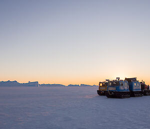 Two Hägglunds oversnow vehicles parked together on the sea ice icebergs in background