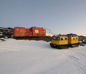 Yellow Hägglunds parked alongside a red field hut