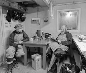 Black and white photo of two expeditioners sitting at a table in a hut