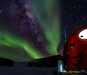 Red round hut with smiley face painted on with an aurora overhead