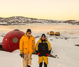 Two expeditioners standing next to a red round fibreglass hut, frozen lake in the background