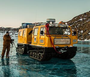 Expeditioner with his camera and tripod standing next to a yellow hagglunds on a frozen lake