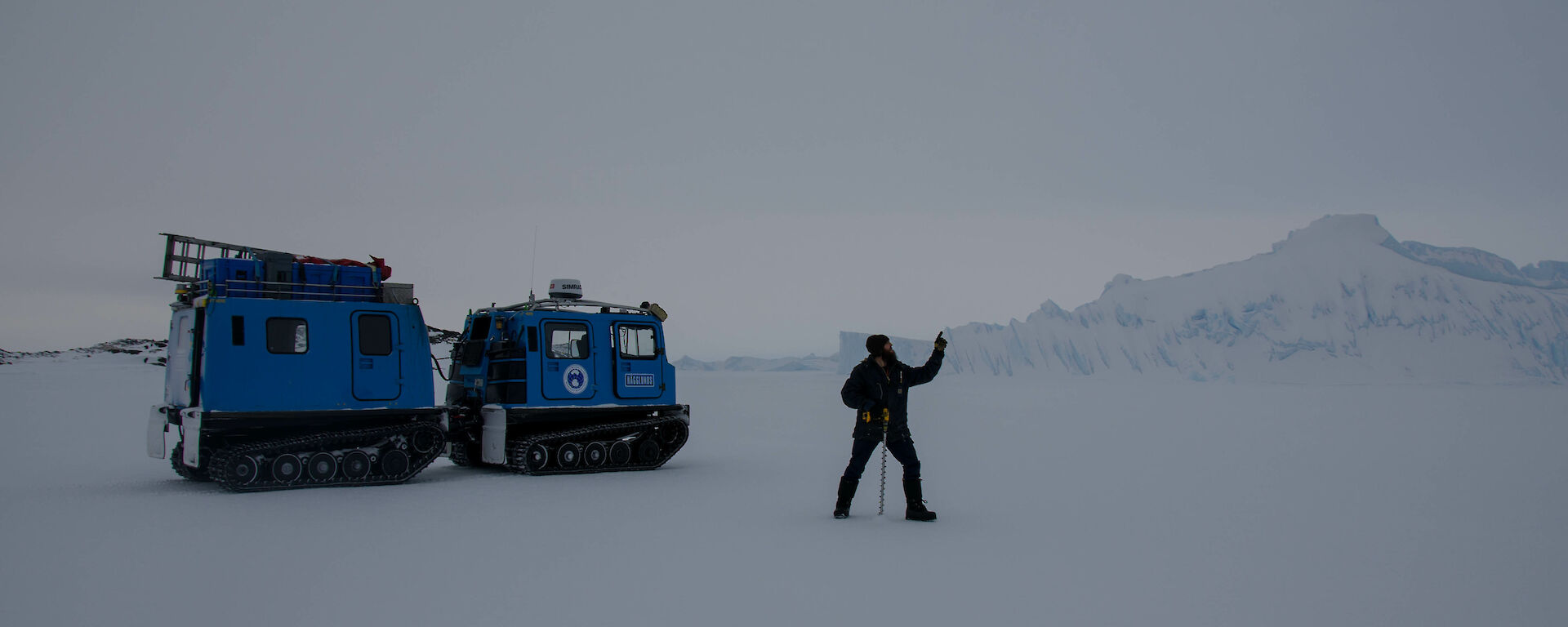 Expeditioner on the sea ice, carrying a sea ice drill, standing next to a blue hagglund