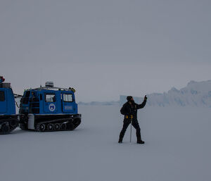 Expeditioner on the sea ice, carrying a sea ice drill, standing next to a blue hagglund