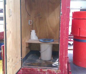 Inside the toilet hut is a white bucket and a toilet seat