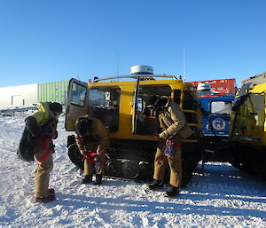 Three expeditioners standing alongside the blue hagglund stepping into harnesses