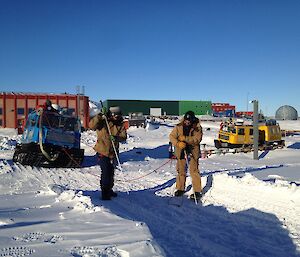 Two expeditioners wearing harnesses attached to a hagglund by rope walking with a large thin stick