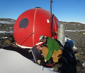 Expeditioner kneeling on the ground alongside a red round hut