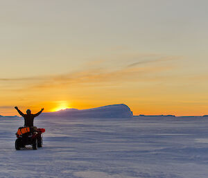 Expeditioner standing on a stationary quad bike with his arms raised sunset in the background