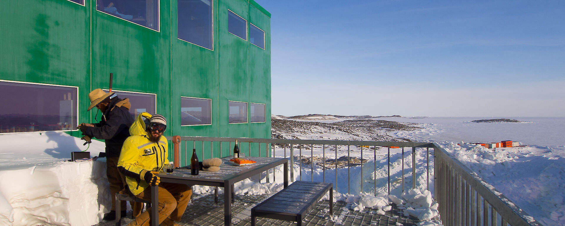 Two expeditioners with Antarctic jackets outside sitting at a table in the sun having drinks
