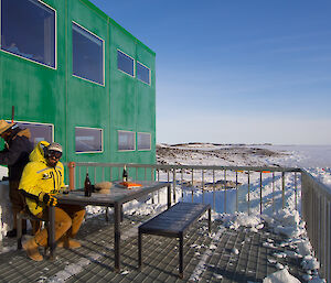Two expeditioners with Antarctic jackets outside sitting at a table in the sun having drinks