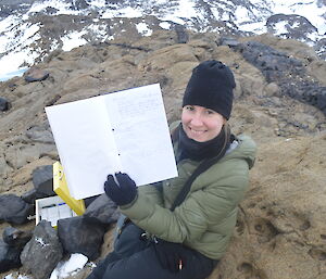 Expeditioner sitting on rocky land holding up a log book