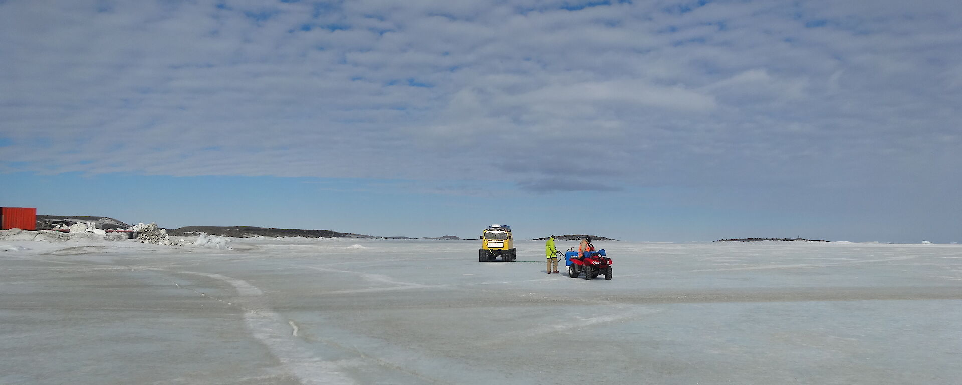 Yellow Hägglunds parked up on the sea ice with a team of 2 attending to it