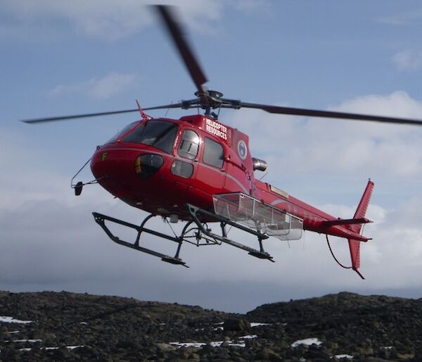 red helicopter with a fitted cargo cage landing with a background of rocky landscape