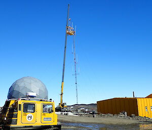 Crane hoisting two communications riggers up to a tower beside the yellow operations building