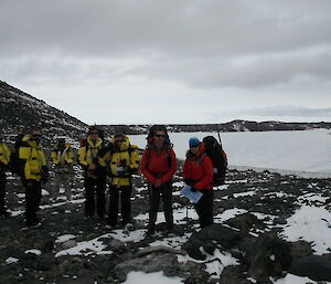 Seven expeditioners in antarctic clothing facing camera against a backdrop of sea ice and rocky landscapes