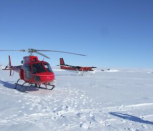 At foreground a red helicopter in front of a twin propeller fixed wing aircraft both on the sea ice