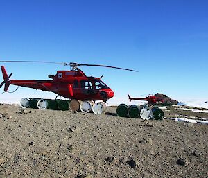 12 fuel drums lying prepared in the foreground next to a red helicopter with another red helicopter in the background.