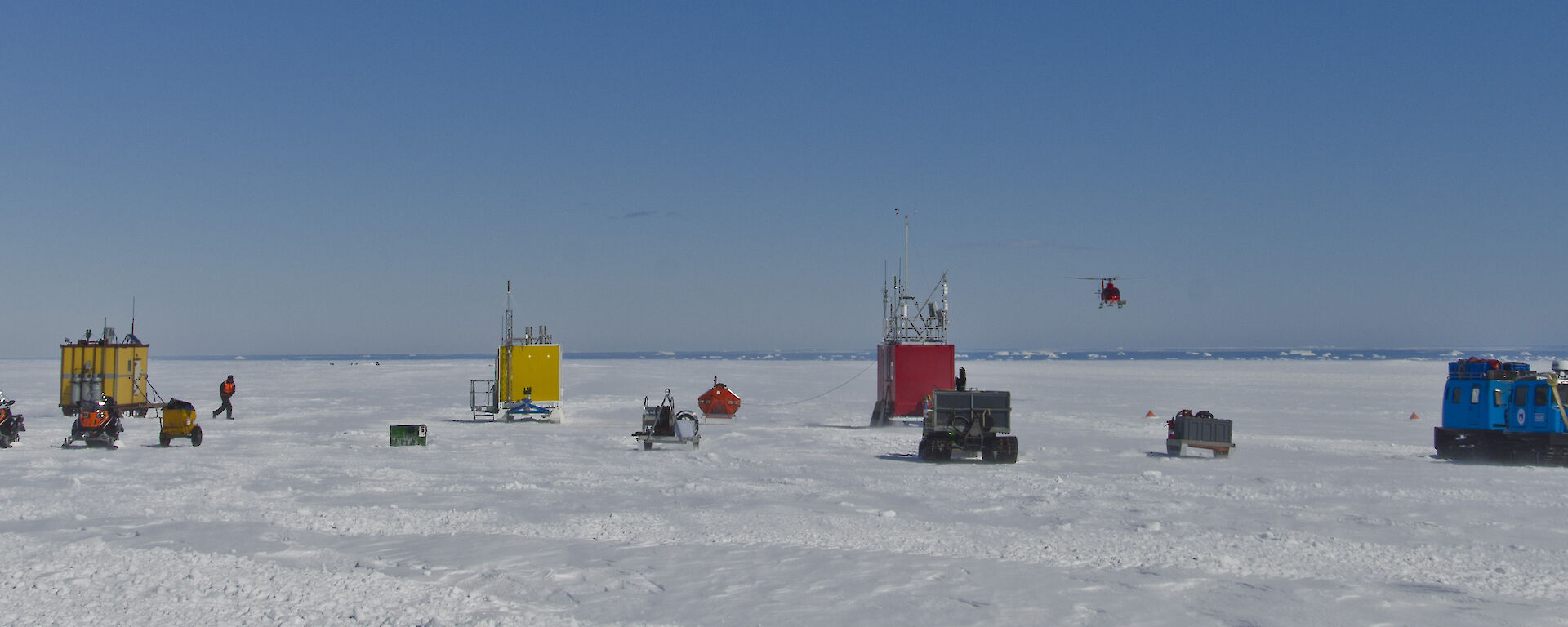 A series of multicoloured prefabricated huts on the plateau at landing site. Helicopter taking off