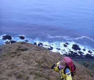 Andrew on the East Coast, photo taken from above as expeditioner climbs up a hill with rocks and sea behind him