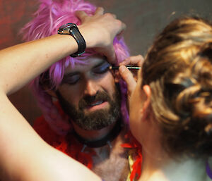 Preparing for the Midwinter play. A male expeditioner in purple wig has eyeshadow applied as part of his costume