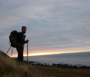 Cameron with hiking gear and poles on a hill with horizon/sunset in background.