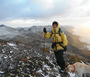 Garry poses with gear on a cliff with sun and sea in background, hunting dog in foreground