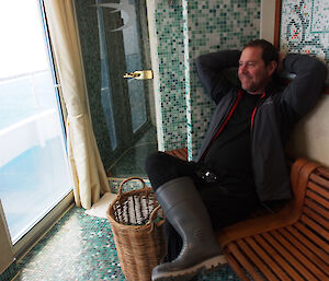 Tom on the Orion posing in a luxurious shower room, sitting on a wooden bench leaning back against a tiled wall