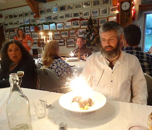 An expeditioner poses with a dessert, in which is a large sparkler which lights his face