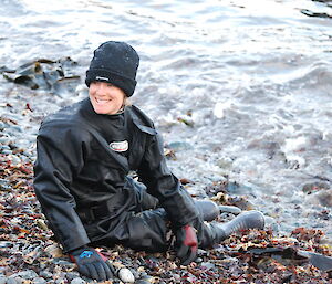 Anna in full dry suit on rocky beach sitting with her toes just out of reach of the tide