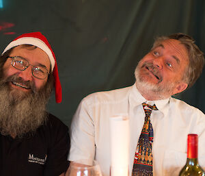 Ray and Jim look amused at Christmas dinner. Ray has a long brown beard and a santa hat while Jim has a short beard that is more grey
