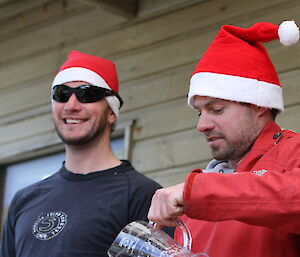 Richard and Andrew smiling, wearing Santa hats and pouring a jug of beer